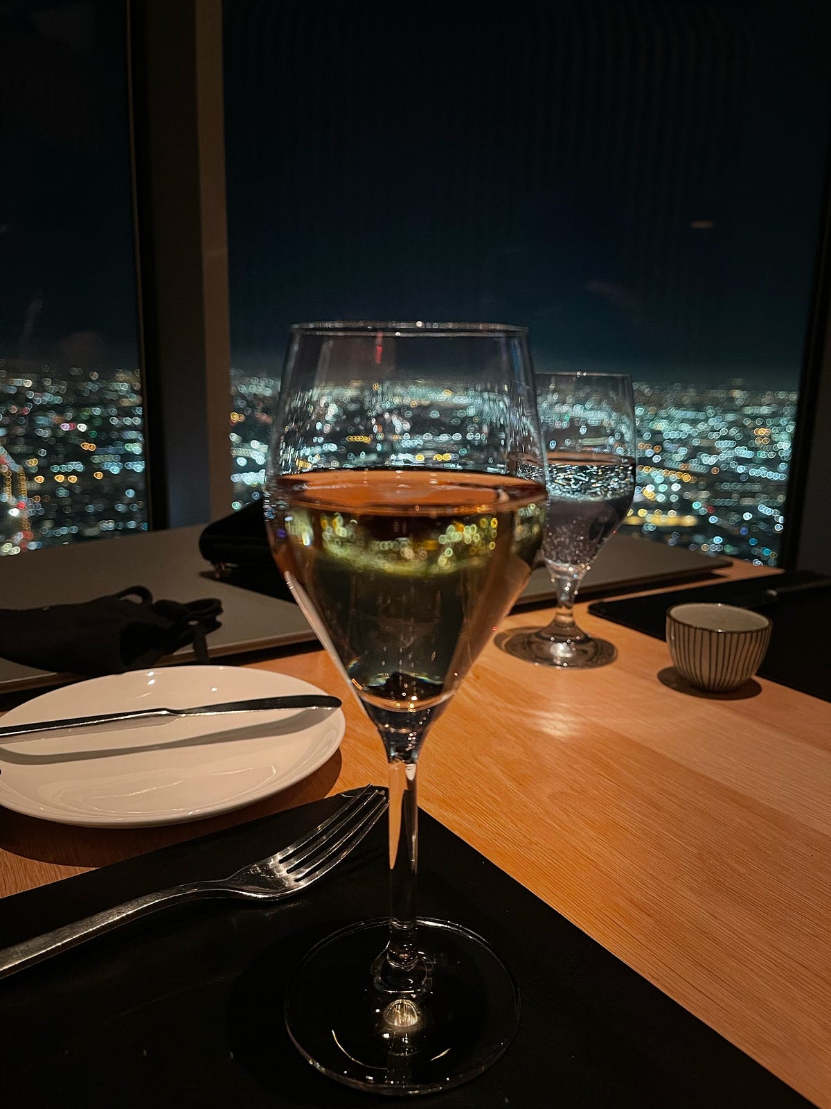 A restaurant table set with wine glasses overlooking a city skyline at night.