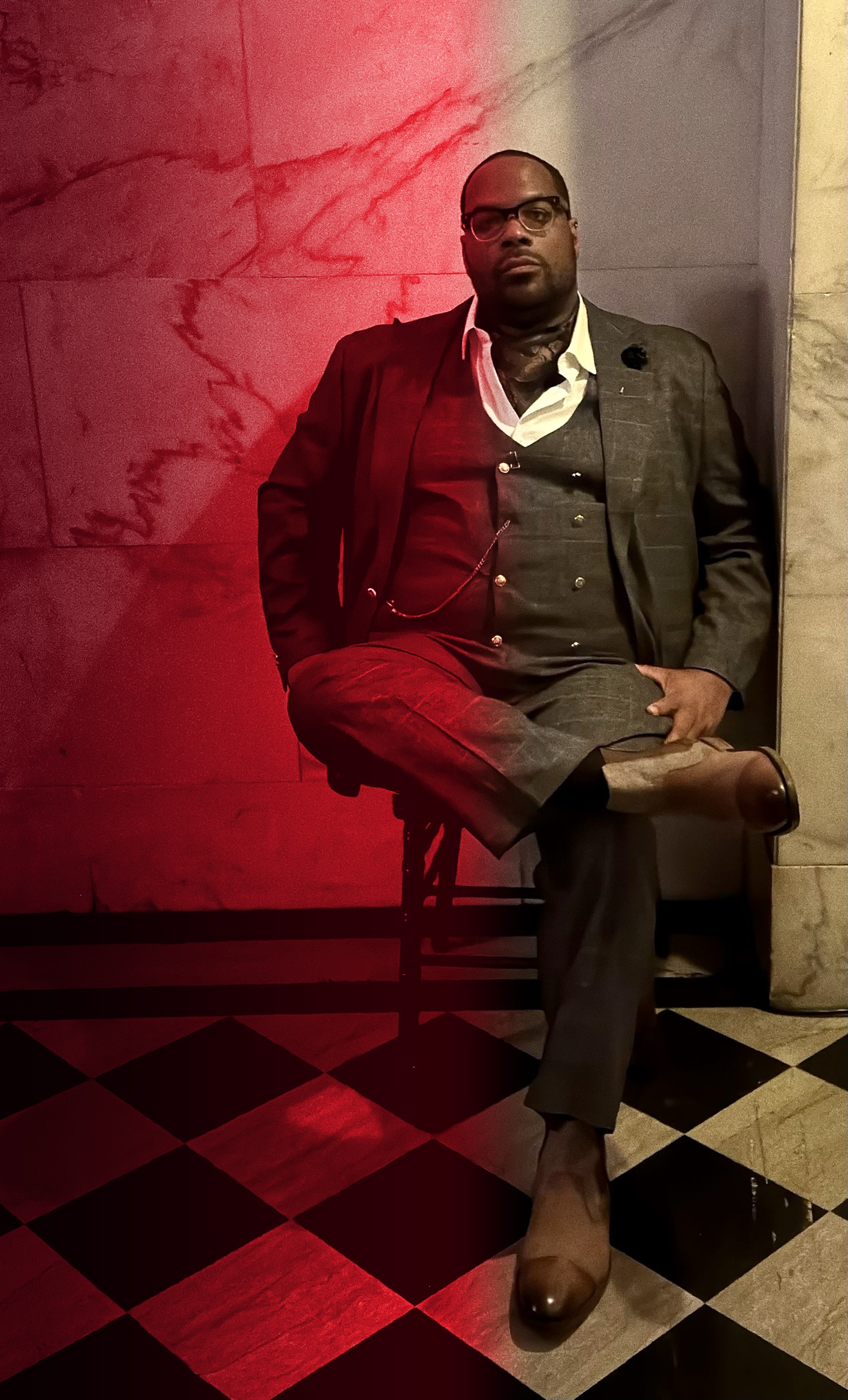 Cue seated in a gray three-piece suit and glasses on a marble floor, the wall behind him lit red on one side.
