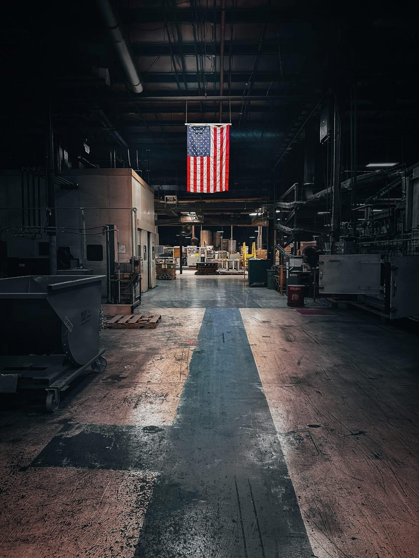 A dimly lit, cavernous factory floor at the FRAM Greenville plant. Heavy industrial machinery and equipment line both sides of a wide concrete aisle marked with a faded blue path. In the center, suspended from the high ceiling, a large American flag hangs brightly illuminated against the surrounding industrial gloom.
